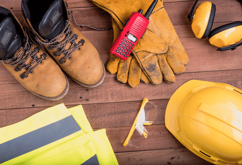 Assorted construction safety gear on a wooden surface, including work boots, gloves, a hard hat, ear protection, safety glasses, a reflective vest, and a red two-way radio.